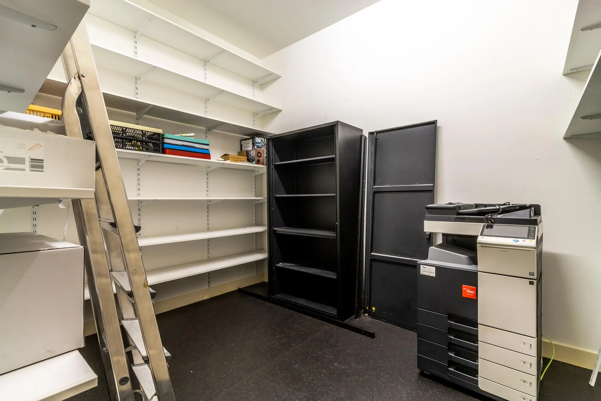 Opbergkamer met metalen trap, witte en zwarte stellingkasten en een groot kopieerapparaat.